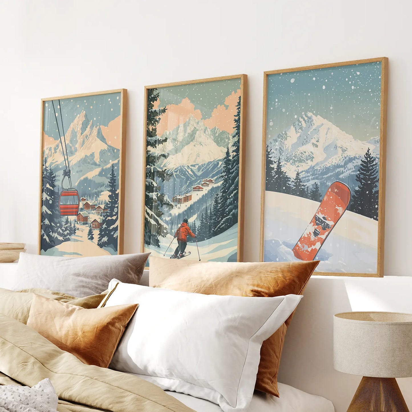 Three framed artworks of a snowy mountain landscape with a skier and snowboard, above a bed with pillows. Natural wood frames