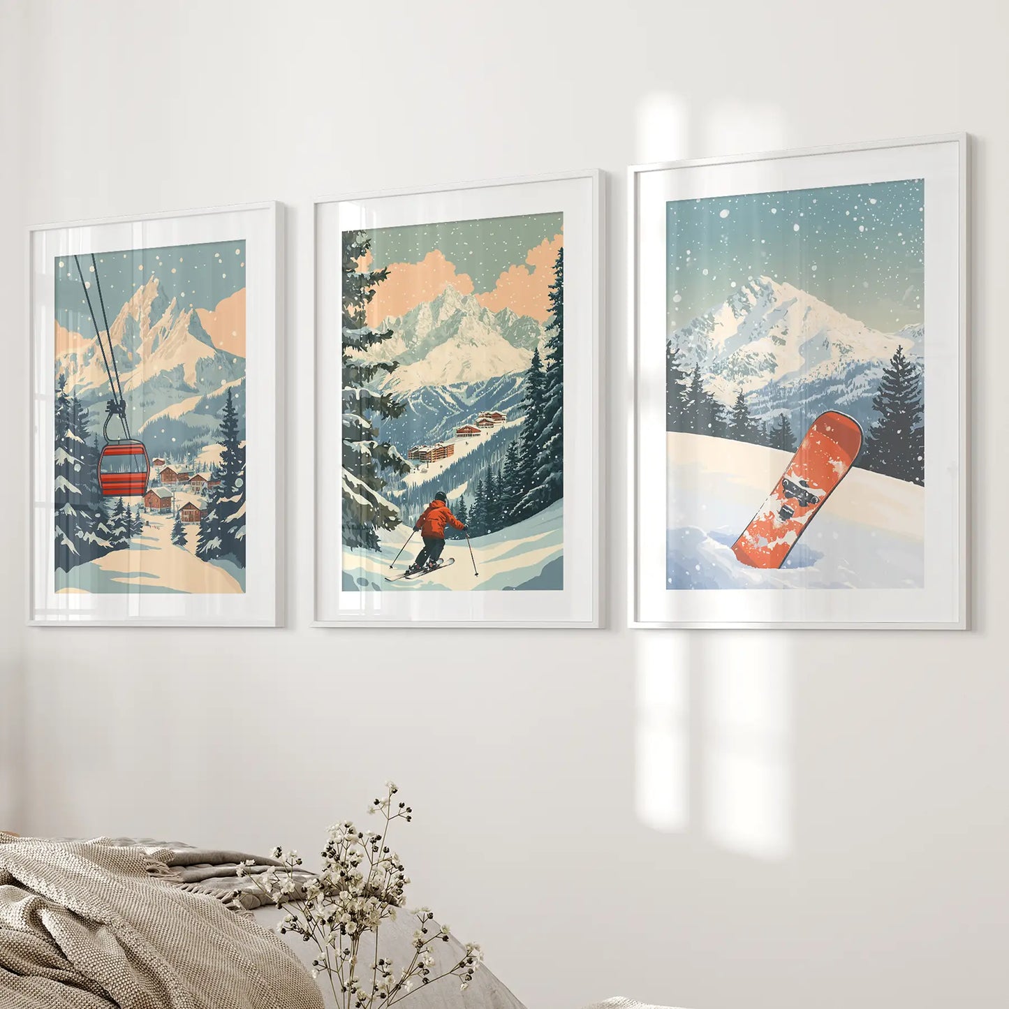 3 framed ski posters. Winter scenes with mountains, a ski lift, and a snowboarder on a white wall above bed. White frames with mat