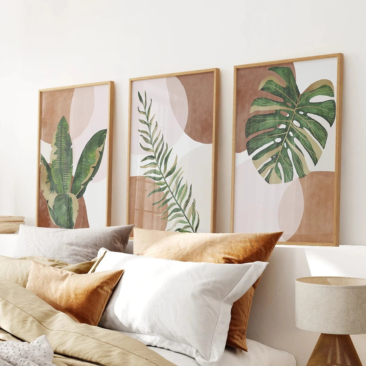 A set of three wood framed prints featuring terracotta backgrounds with abstract botanical green tropical leaves, displayed above a bed with beige and white bedding.