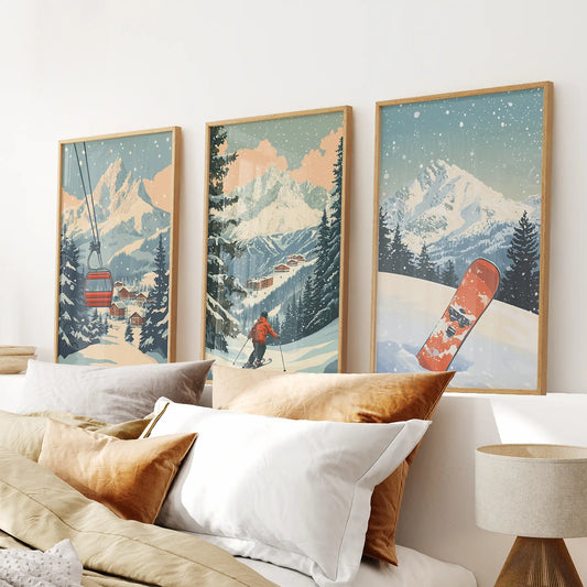 Three framed artworks of a snowy mountain landscape with a skier and snowboard, above a bed with pillows. Natural wood frames