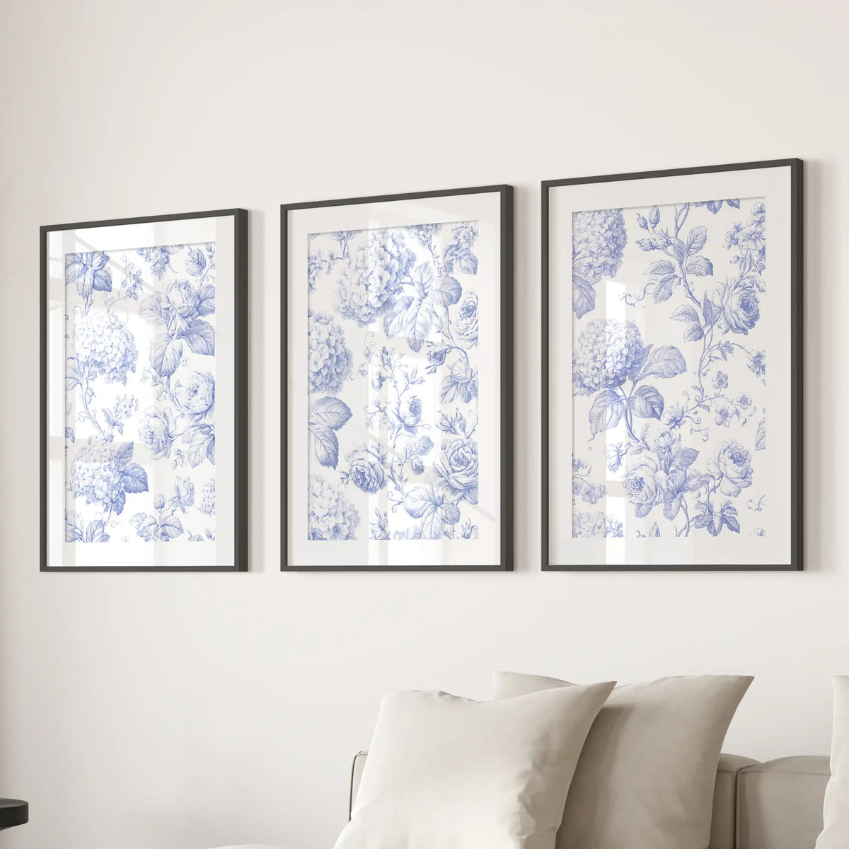 The three matted black frames above a sofa hold blue floral prints, with curling stems and leaves on warm cream backgrounds.