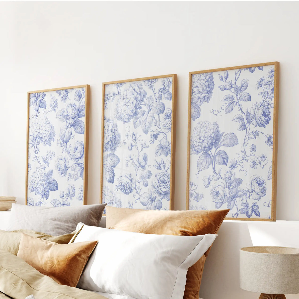 The three oak-framed prints above a bed show blue flower heads among curling stems, with cream backgrounds visible between the leaves.