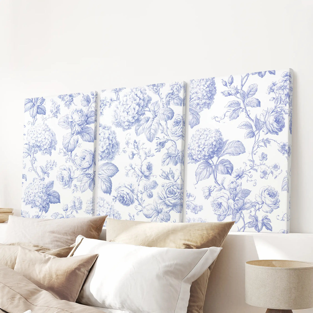 Blue roses, hydrangea heads and curling leaves fill three separate canvases above the bed pillows, against warm cream backgrounds.