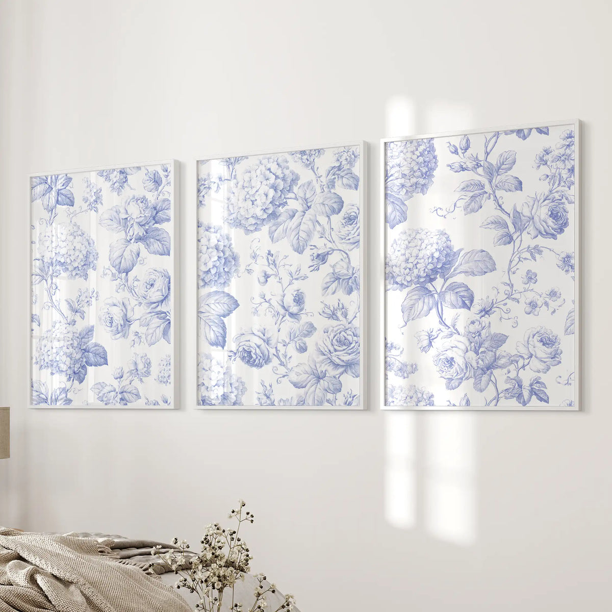 A blue floral pattern of roses, rounded flower clusters and fine leaves spans three white-framed prints beside a bed.