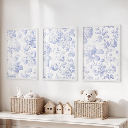 A set of three prints in white frames shows blue flowers and fine leaves against warm white above a toy storage bench.