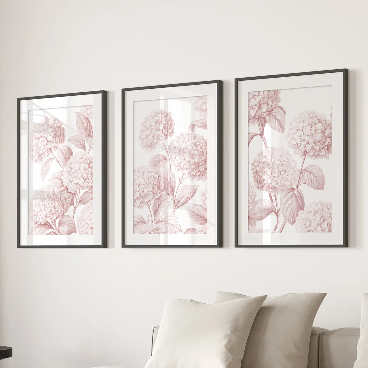 Fine pink outlines trace clusters of hydrangea flowers and branching leaves in three matted black frames above a sofa.