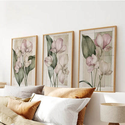 Long green leaves and pale pink blossoms fill three oak-framed prints above white and ochre bed pillows, with faint rectangular lines behind them.