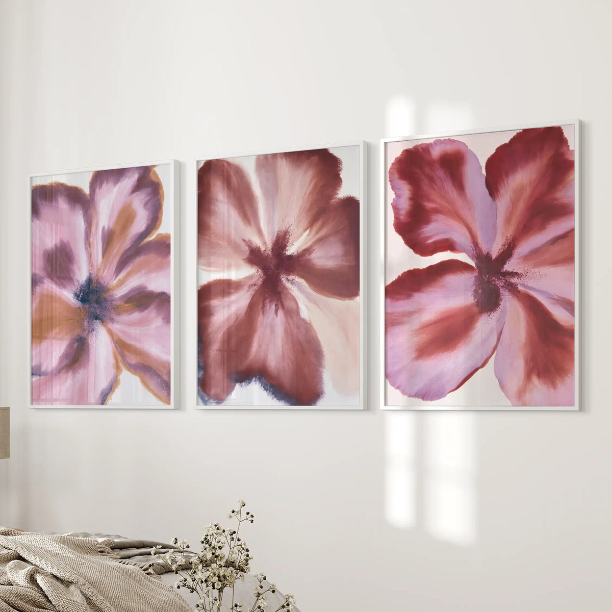 A set of three white-framed prints beside a bed shows close views of flowers, with wine-red petals fading into dusty pink.
