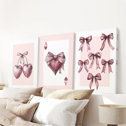 Unframed canvas pieces show the coquette pink icons-ace, bows, and cherries-stacked above the headboard as a lively three-piece wall art display.