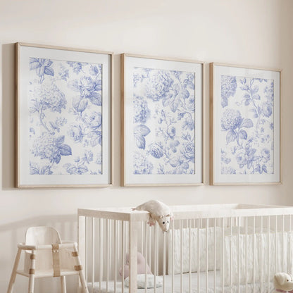 Above a crib hangs a set of three prints showing blue hydrangea clusters and roses on cream backgrounds, framed in oak with white mats.