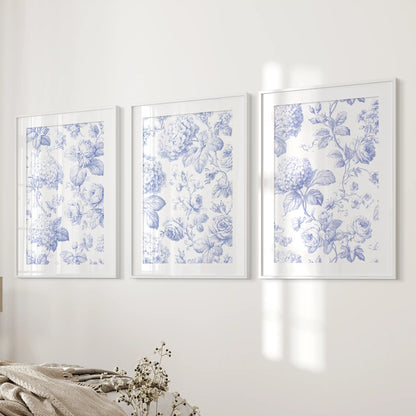 Beside a bed, three prints in white frames with mats show blue hydrangea clusters and roses on cream backgrounds.