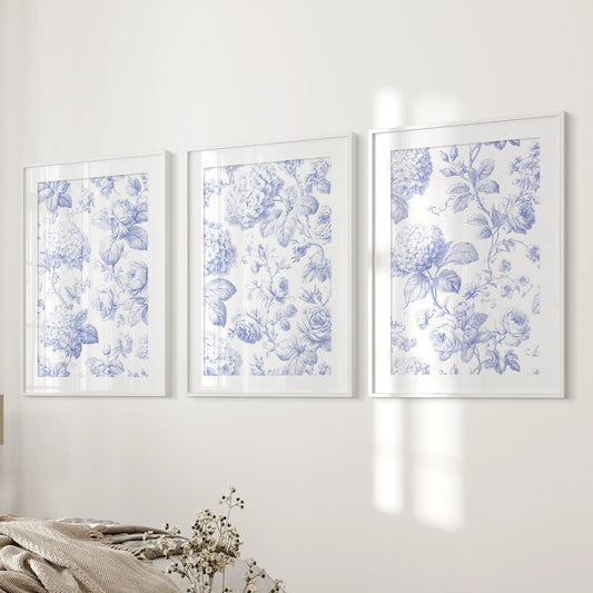 Beside a bed, three prints in white frames with mats show blue hydrangea clusters and roses on cream backgrounds.