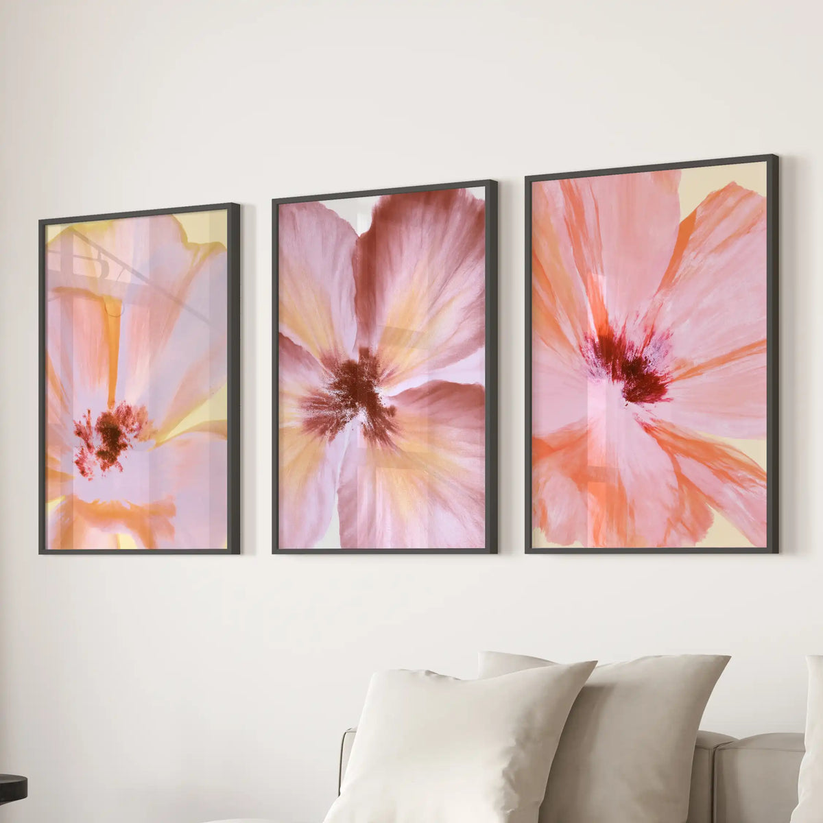 Peach and pink petals overlap in three black-framed flower prints above a sofa, with dark red centers visible among the broad strokes.