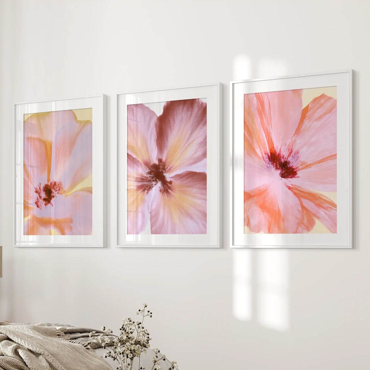 White mats and frames surround three flower prints beside a bed, showing overlapping peach and pink petals around dark red centers.
