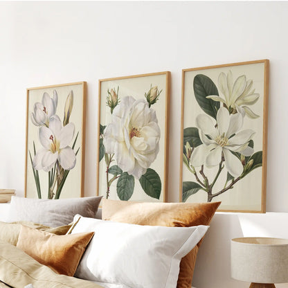 Above a bed, three oak-framed prints display white flowers, including a rose.