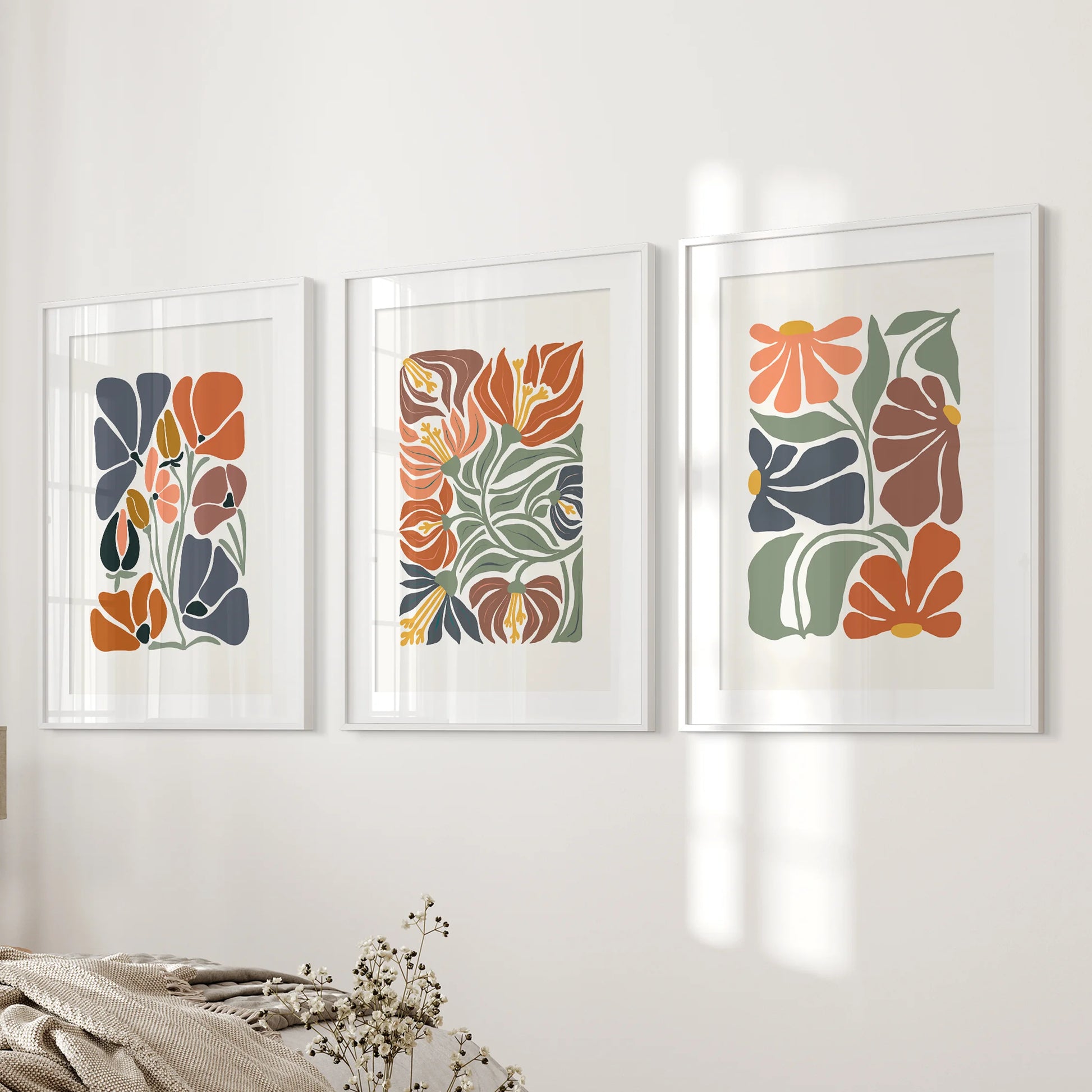 Matted white frames hold three floral artworks that soften the bedside wall with gentle blooms and grounded terracotta color.