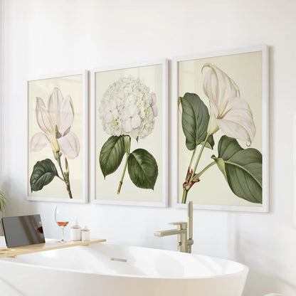 White frames surround three botanical prints above a bathtub, with a central hydrangea head and larger white blossoms on either side.