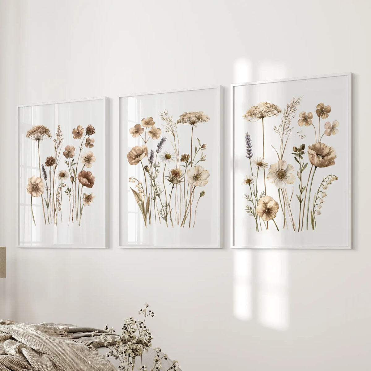 Three prints of slender meadow stems with beige and brown flowers hang beside a bed in white frames.