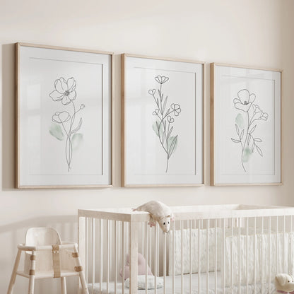 A trio of minimal floral prints in wood-matted frames hangs above the infant bassinet, soft grain framing the clean botanical lines.