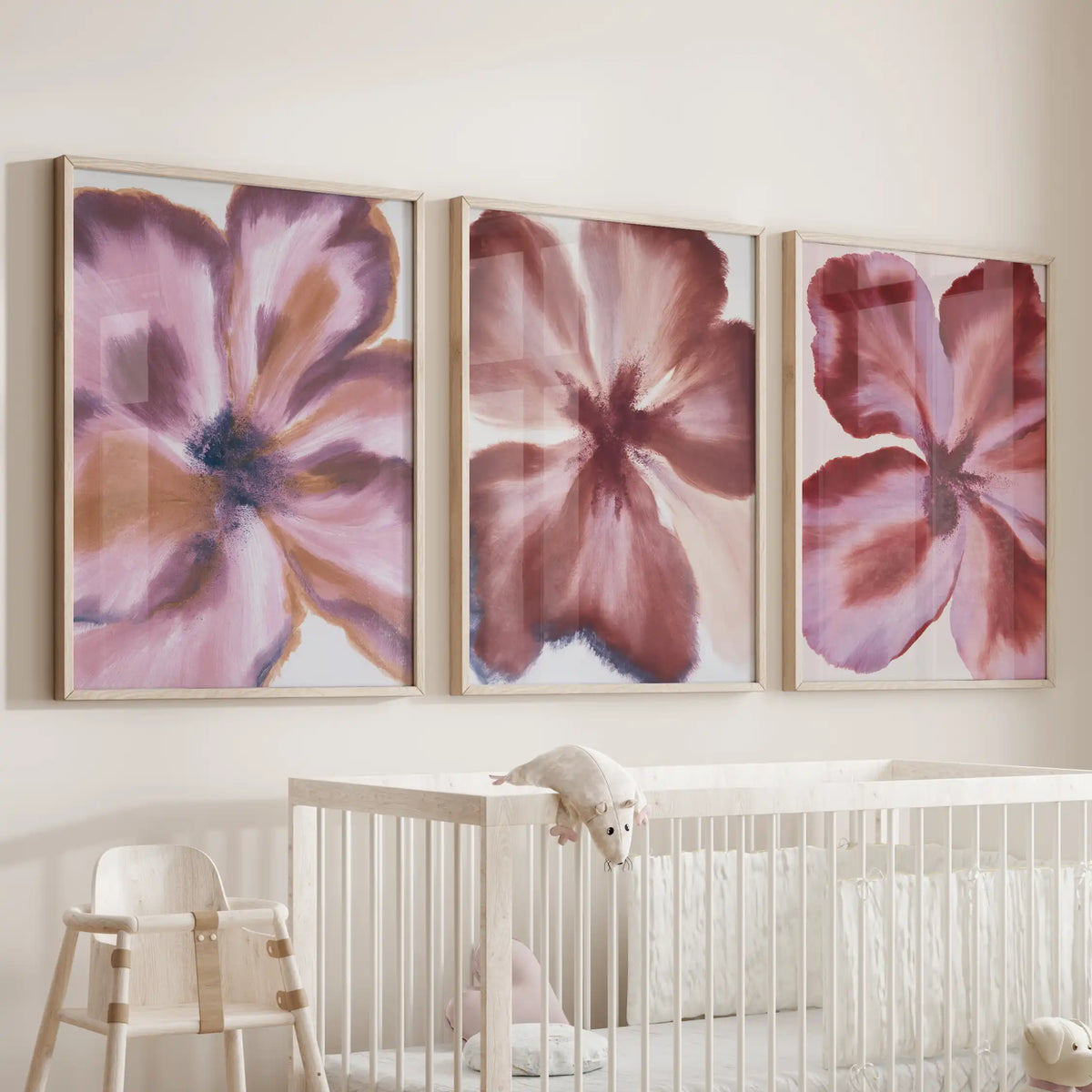 A set of three pink and wine-red floral prints above a nursery crib, in oak frames.
