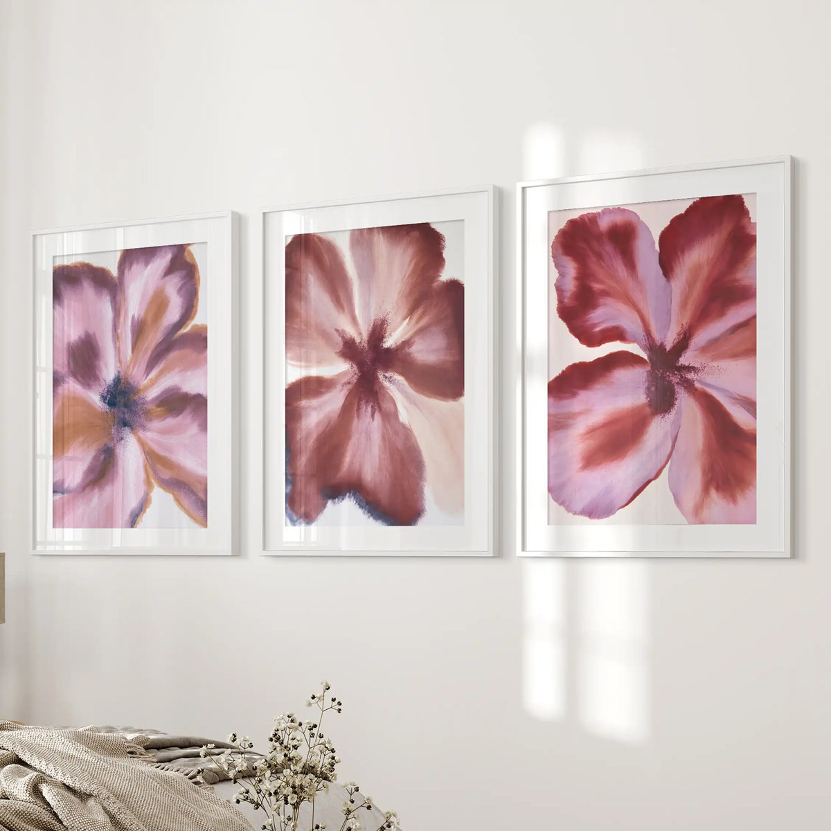 White mats border broad wine-red and mauve petals in three white-framed prints beside a bed, with dark centers visible among the strokes.