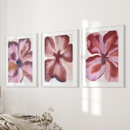 White mats border broad wine-red and mauve petals in three white-framed prints beside a bed, with dark centers visible among the strokes.