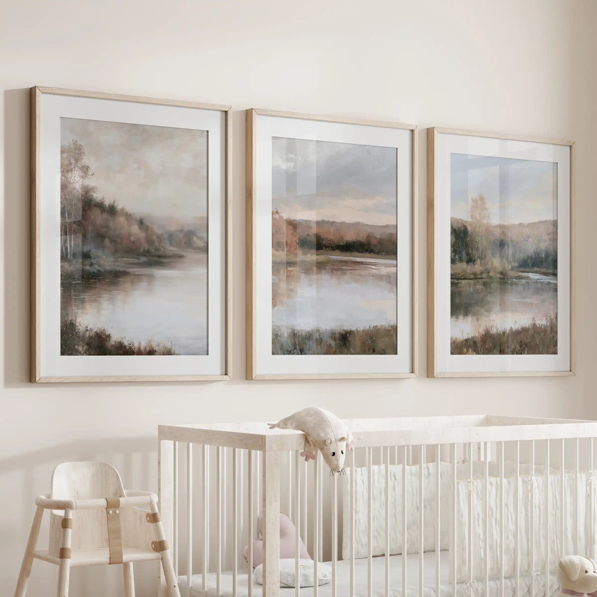 A set of three nature art prints featuring a cottage lake house hangs over the bassinet, matted in oak frames with gentle, earthy tones.