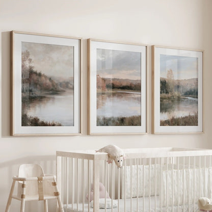 A set of three nature art prints featuring a cottage lake house hangs over the bassinet, matted in oak frames with gentle, earthy tones.