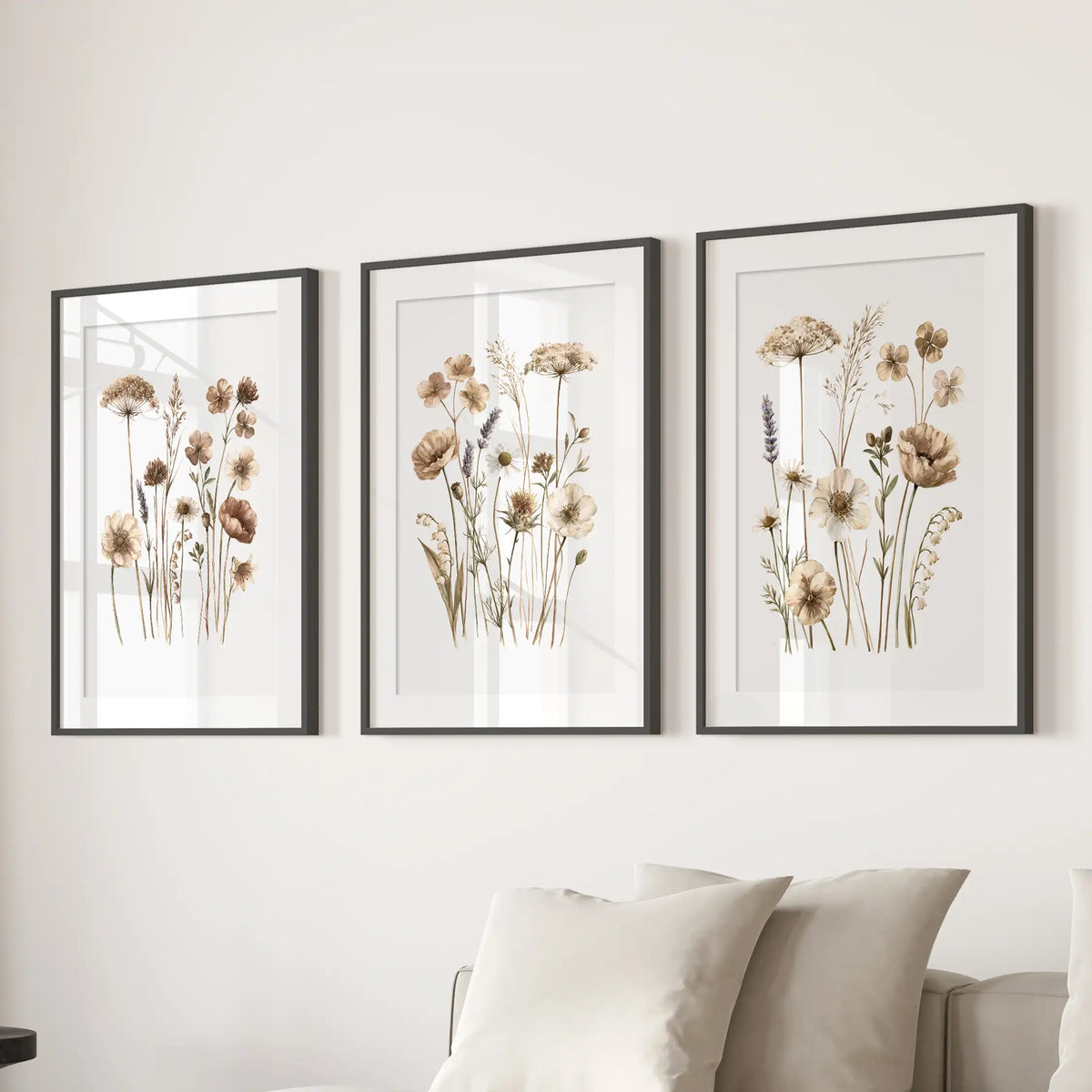 The three black frames above a sofa have white mats around delicate meadow stems with small beige flowers on white backgrounds.