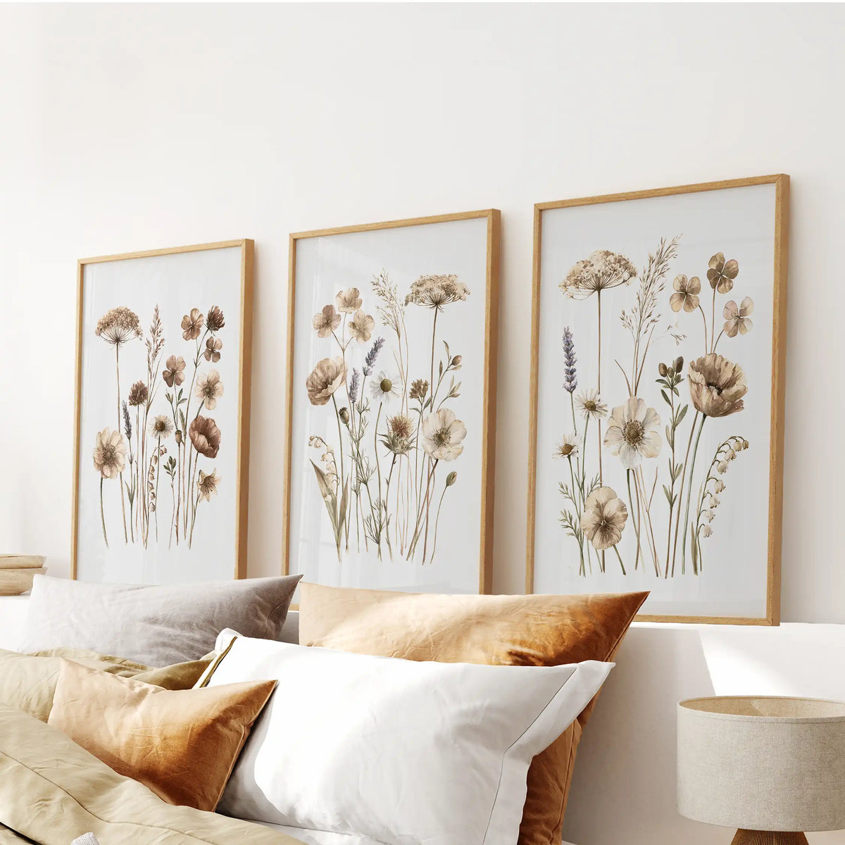 A set of three neutral wildflower prints in oak frames above a bed.