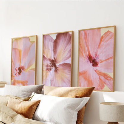 The dark red centers and broad pink petals of three flower close-ups appear in oak frames above white and ochre bed pillows.