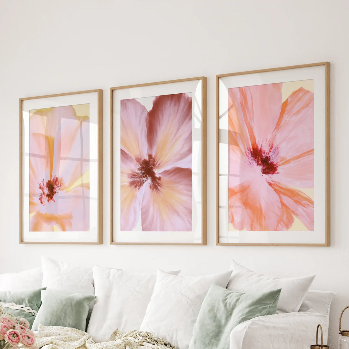 A set of three pink and peach flower prints in oak frames with white mats above a sofa.
