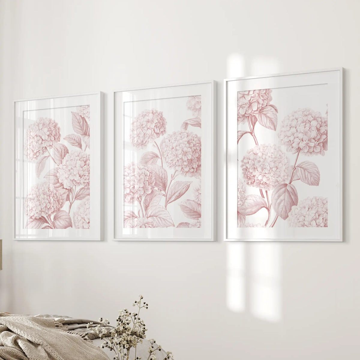 Beside a bed, white frames and mats surround three pale pink hydrangea patterns with fine leaf and petal outlines.