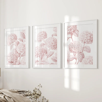 Beside a bed, white frames and mats surround three pale pink hydrangea patterns with fine leaf and petal outlines.