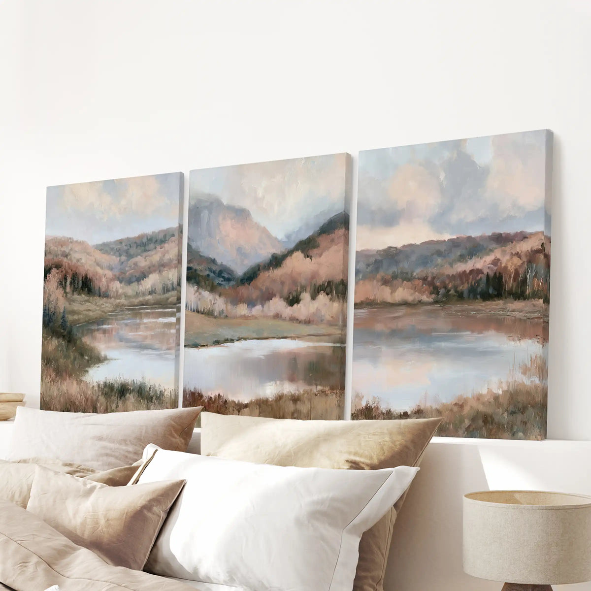 A three-panel canvas wall art set depicting a calm lake and forested hills under a cloudy sky, displayed above a bed with neutral-toned pillows in a softly lit bedroom.