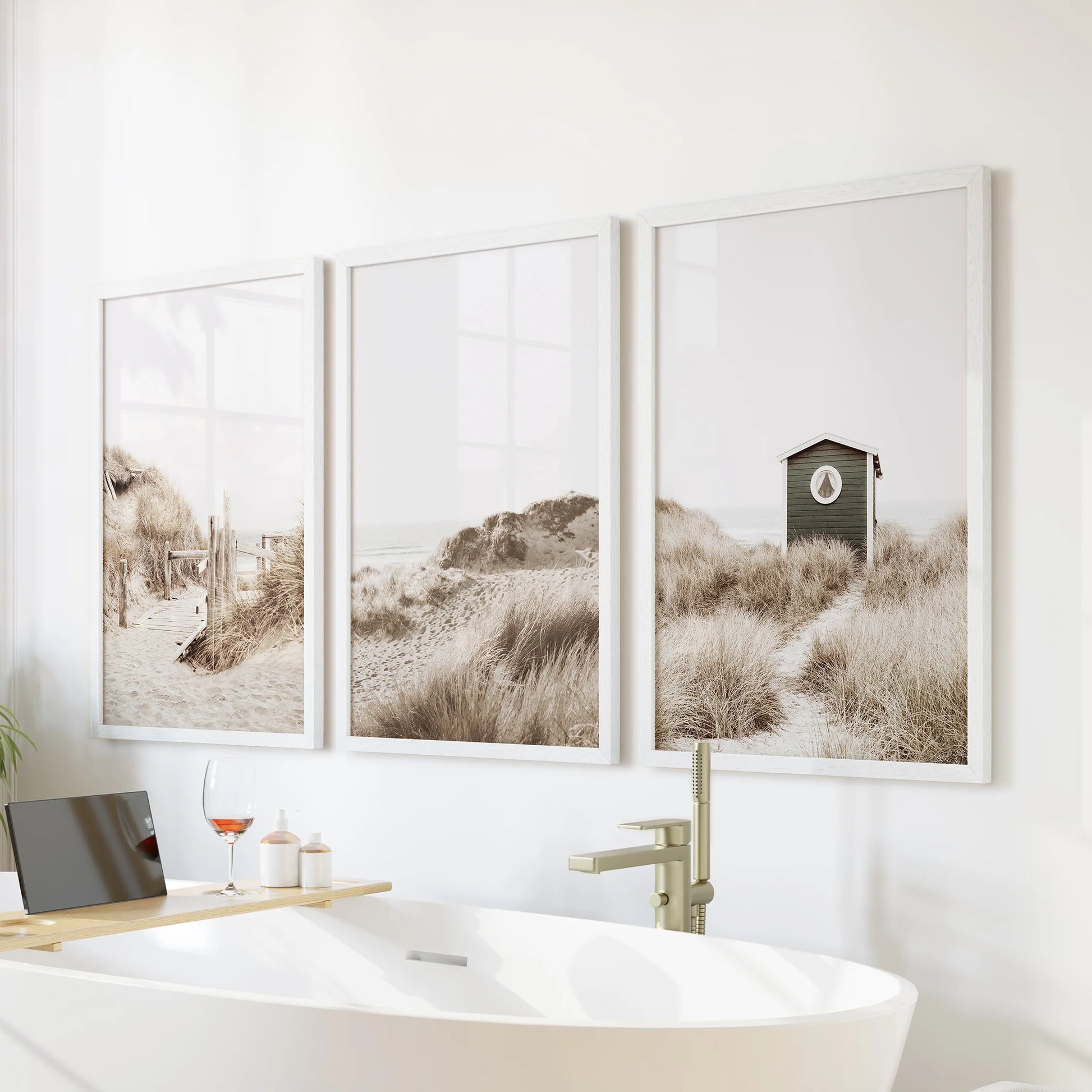A white-framed coastal trio hangs over the bathtub wall, soft dunes and pastel sky giving the bathroom a spa-like, serene feel.