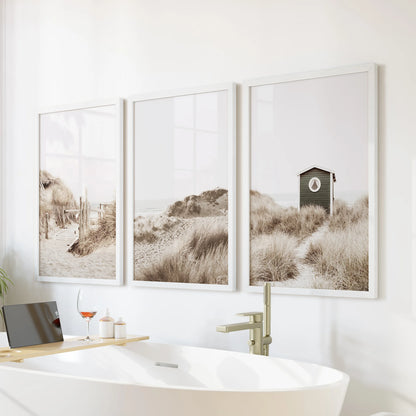 A white-framed coastal trio hangs over the bathtub wall, soft dunes and pastel sky giving the bathroom a spa-like, serene feel.