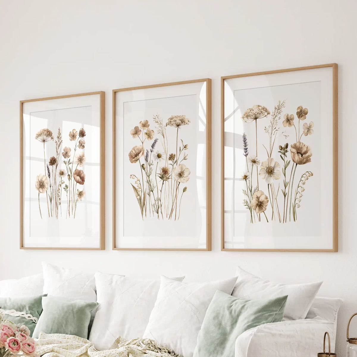 Small meadow blooms on slender stems fill three prints above a sofa, with oak frames and white mats around their white backgrounds.