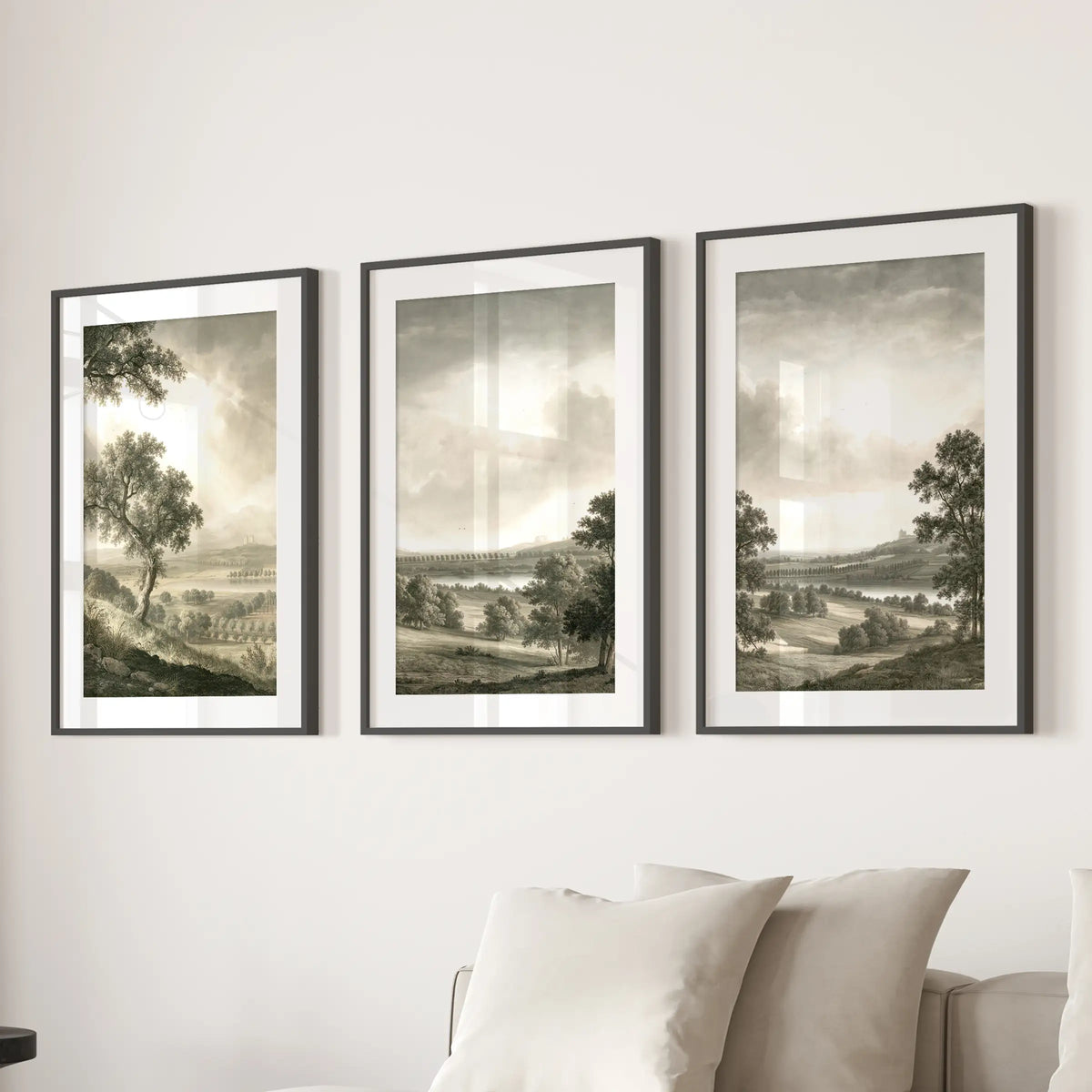 Rural landscape wall decor in three pieces, shown in black frames with white mats above a sectional sofa in a lounge.