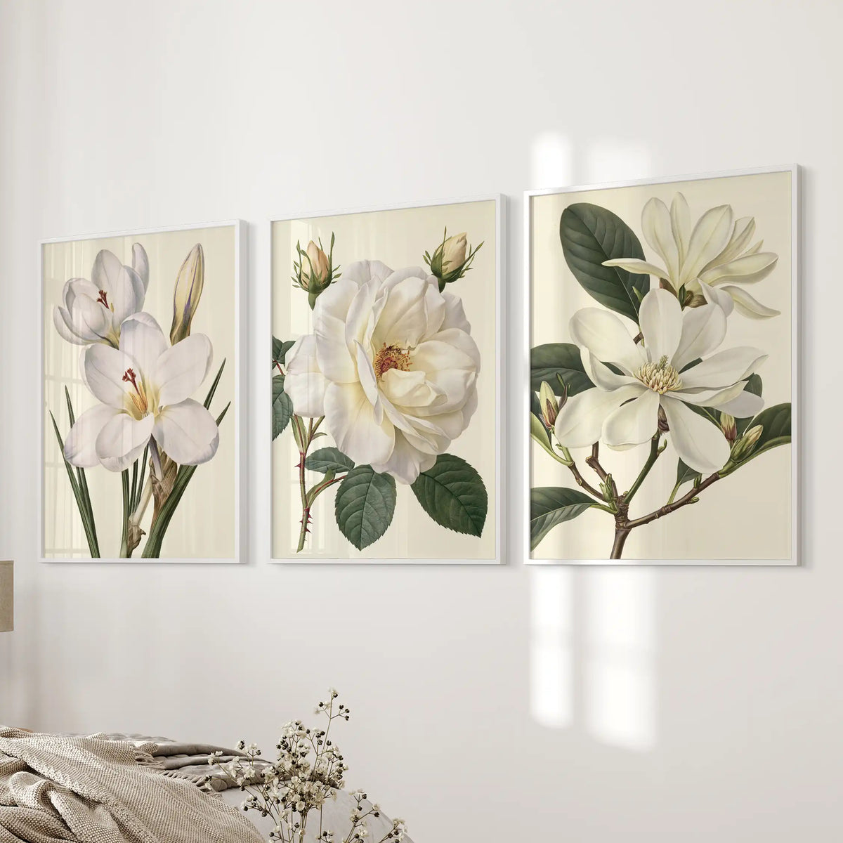 Three white frames hold prints of white petals and green leaves on cream backgrounds beside a bed.