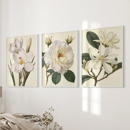 Three white frames hold prints of white petals and green leaves on cream backgrounds beside a bed.