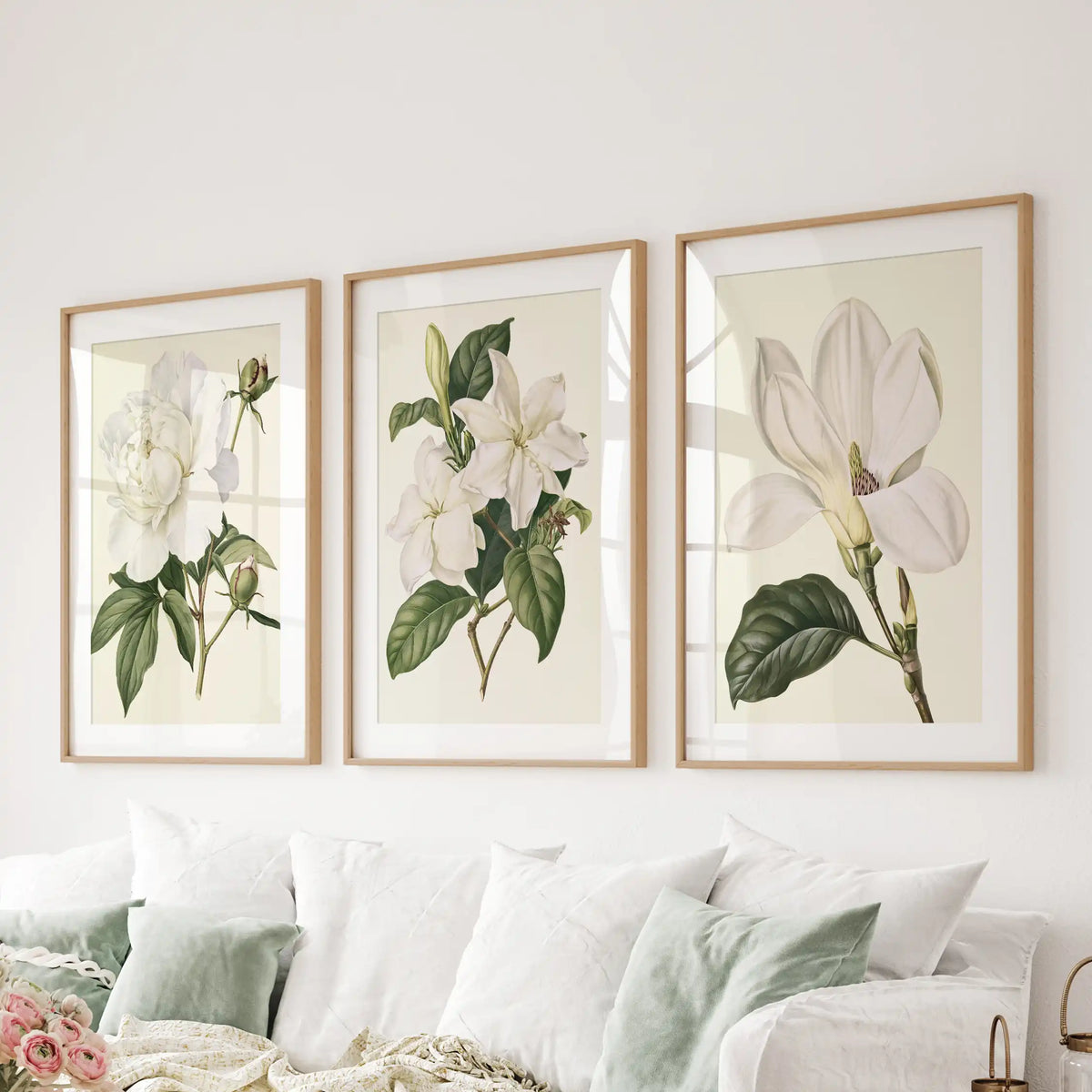 White mats surround three cream-backed floral studies above a sofa, with ruffled petals, small blossoms and a large open flower in oak frames.