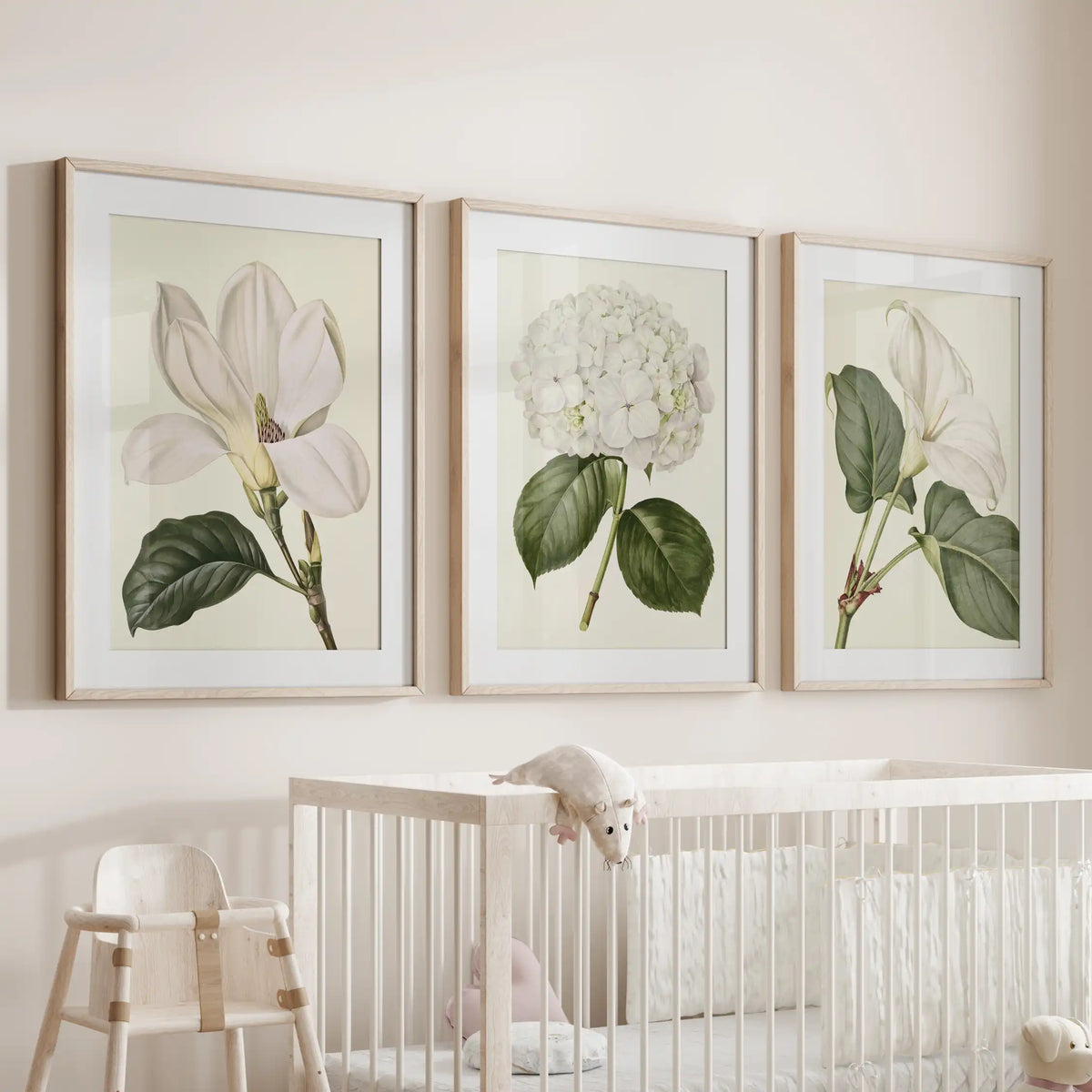 Above a white crib, three matted oak frames show a central hydrangea head between larger white flowers with broad green leaves.