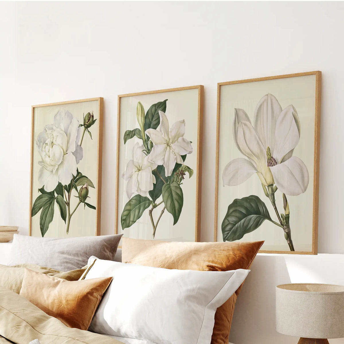 Above the bed pillows, three oak-framed prints show white blooms and dark leaves on cream, with round buds beside the left-hand flower.